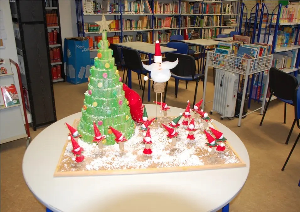 Árvore de Natal verde rodeada de pequenos duendes de gorro vermelho, sobre uma mesa redonda de uma biblioteca escolar