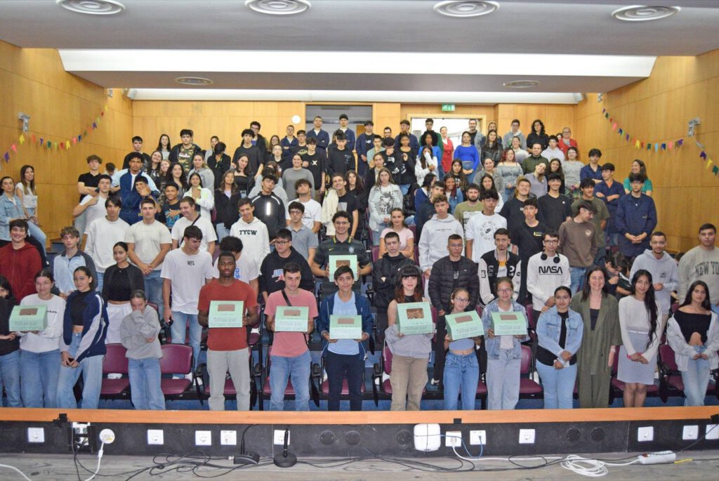 Plateia de alunos e professores num auditório, com várias pessoas na primeira fila a segurar placas verdes