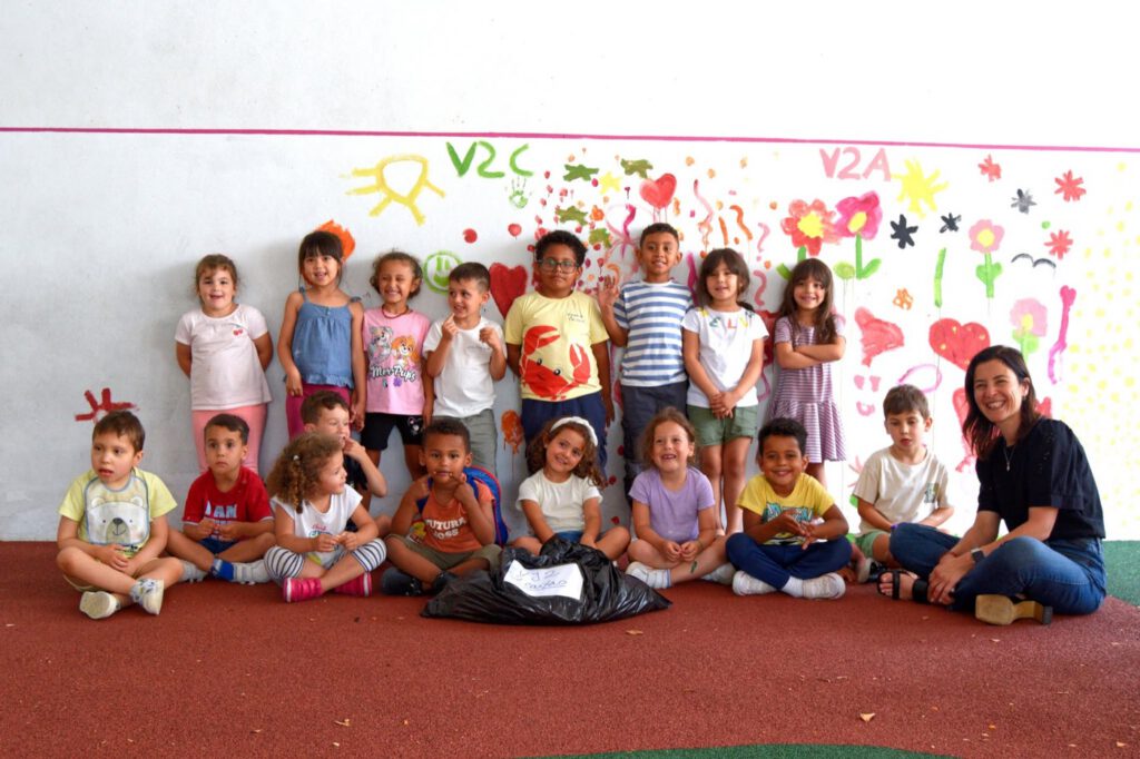 Crianças pequenas com uma educadora, numa sala, em frente a uma parede pintada com flores