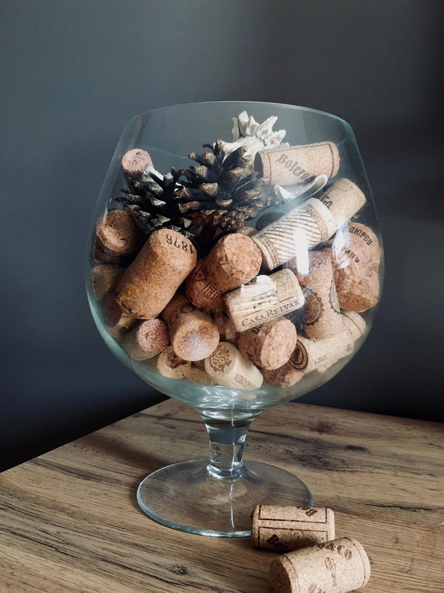Taça de vidro cheia de rolhas de cortiça e pinhas, sobre uma mesa de madeira
